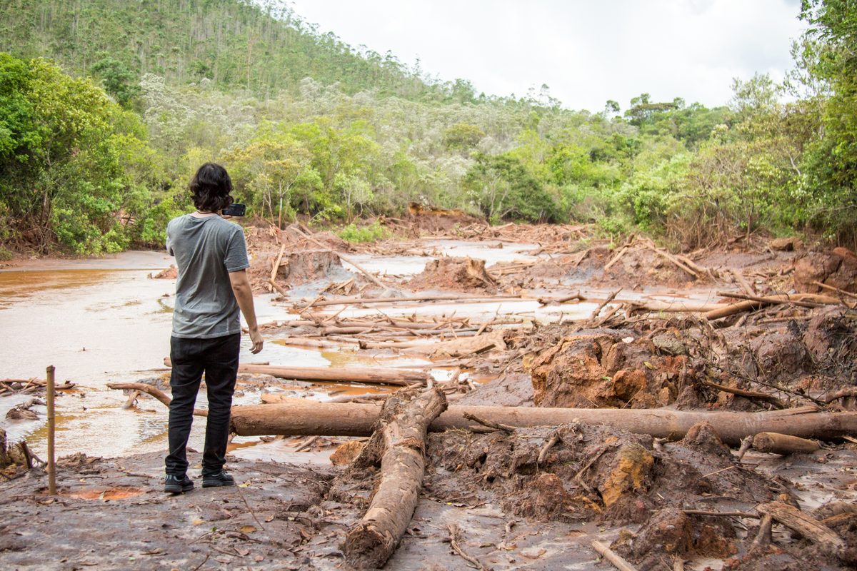 Um homem observa o leito de um rio tomado por lama e destroços de árvores após o rompimento da barragem.